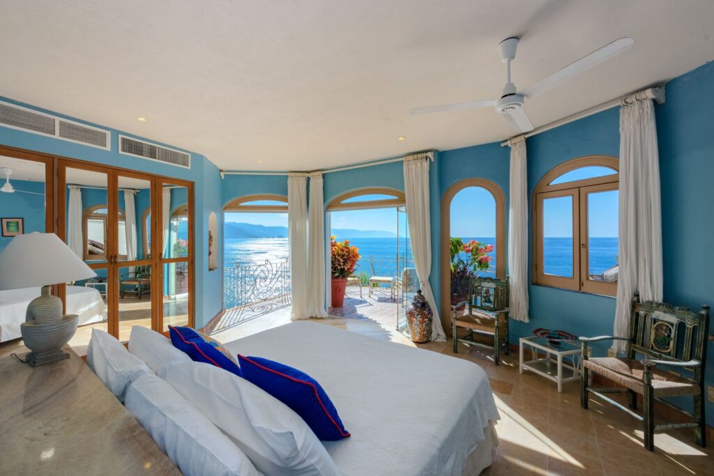 Bright seaside hotel room with white bedding, blue walls, and large arched balcony windows overlooking the ocean