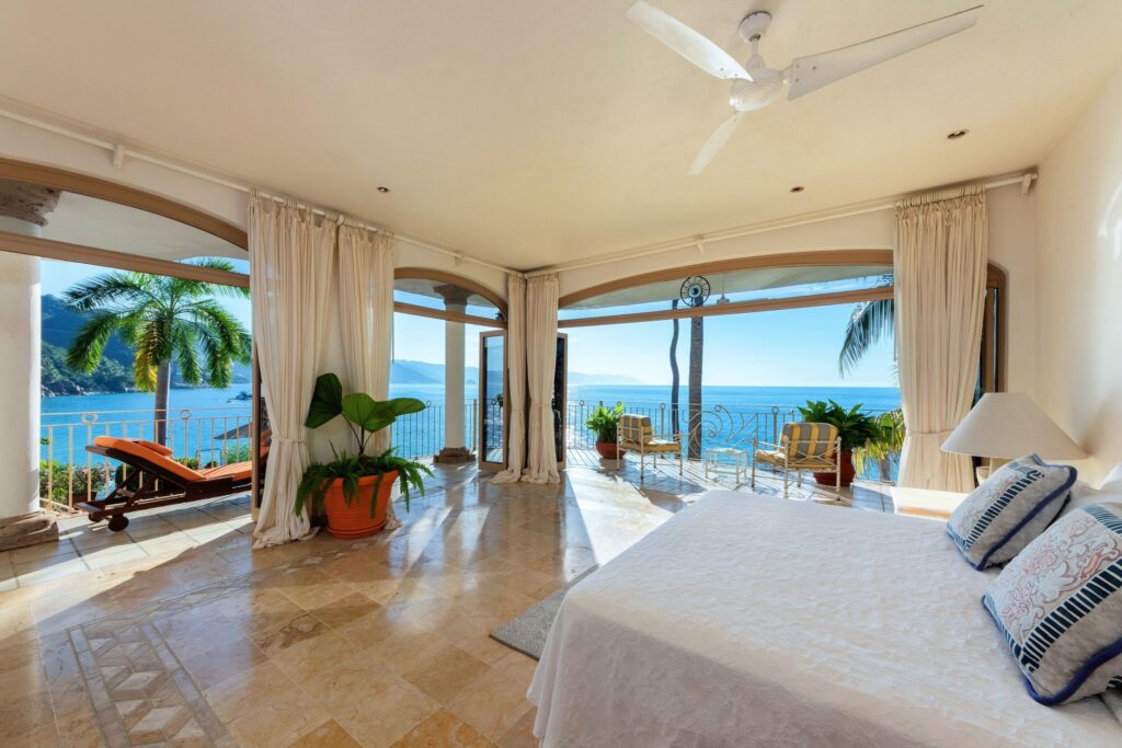 Luxurious bedroom with white linens and ceiling fan, opening to an oceanfront balcony with palm trees and blue sea beyond.