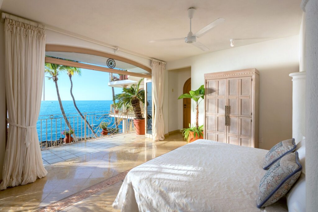 Bright hotel bedroom with white bedding, beige curtains, and a wooden armoire; balcony doors reveal palm trees and blue ocean beyond