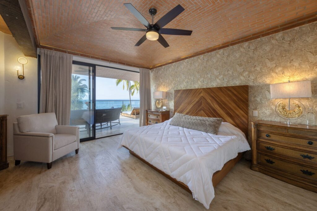 Hotel bedroom with white bed, wooden headboard, stone wall, ceiling fan, and ocean-view balcony through glass doors.