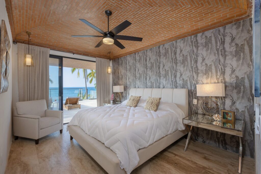 Bedroom with white bed and beige seating, sliding doors to a balcony overlooking the ocean; brick vaulted ceiling and textured gray wall visible on the right.”,
