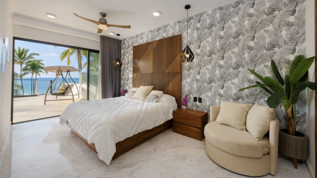 Hotel bedroom with white comforter, wooden headboard, and tropical wallpaper overlooking a balcony with an ocean view.