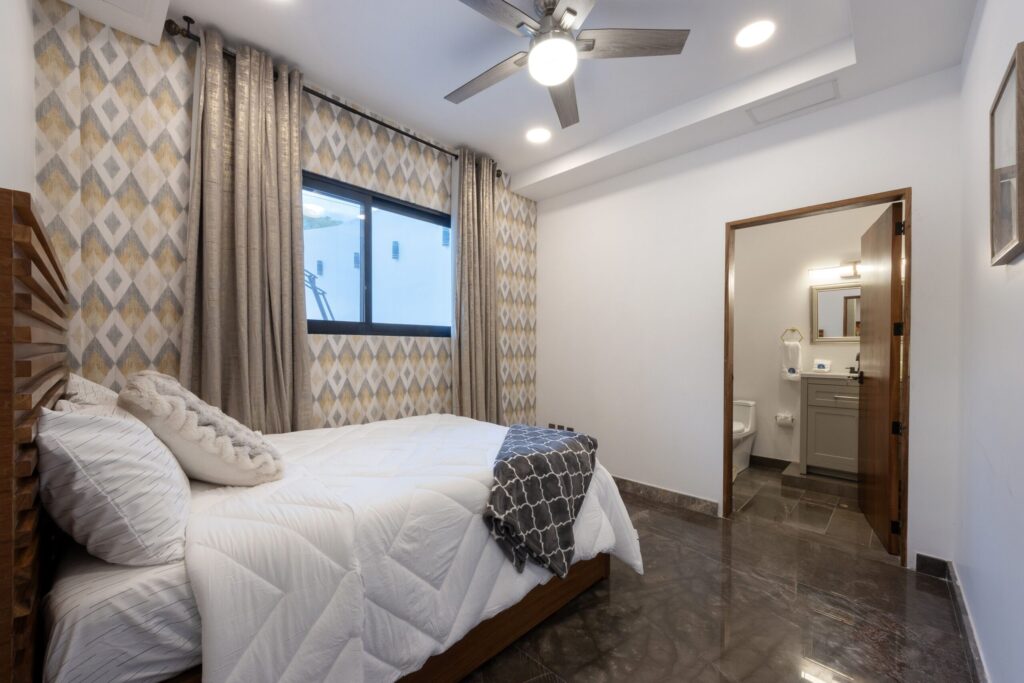 Bedroom with two white-quilted beds and a geometric wallpaper accent wall, beige curtains, and a ceiling fan; open bathroom visible to the right.