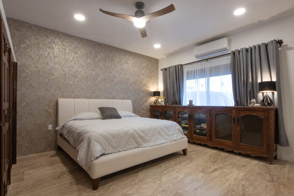 Bedroom with beige upholstered bed, textured accent wall, and wooden dresser against a windowed wall with gray curtains