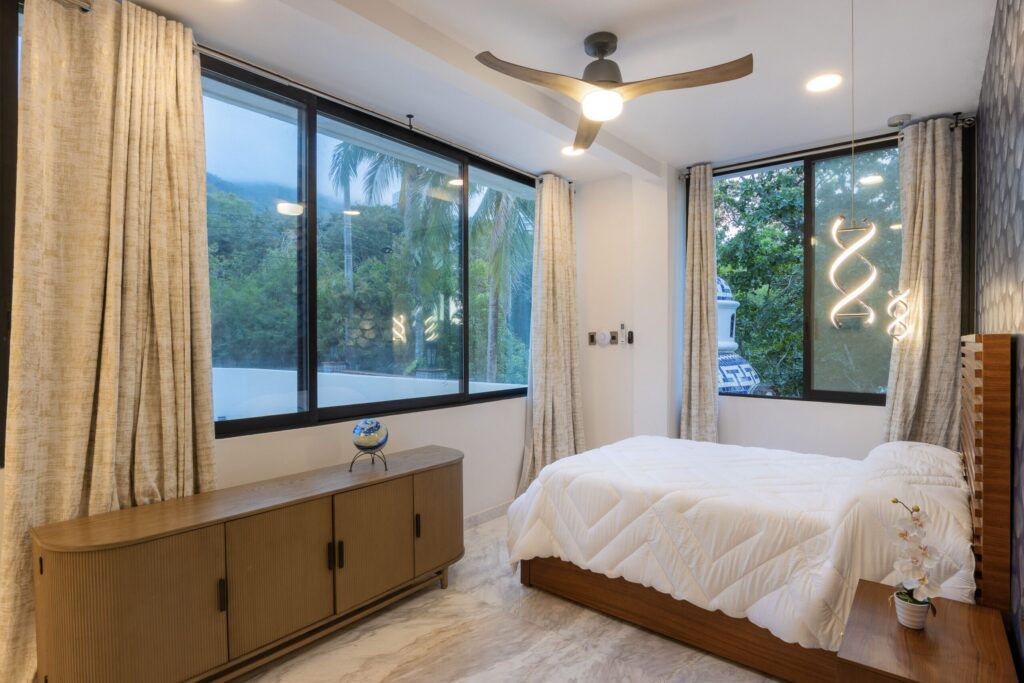 Bright modern bedroom with large windows and beige curtains, white quilted bed, and a wooden console along the wall beneath the windows.