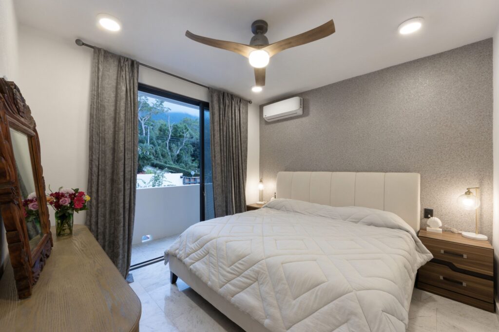 Twin bed bedroom with white quilts, wood nightstands, ceiling fan, and large window with gray curtains overlooking greenery