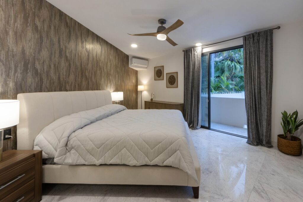 Modern bedroom with a white upholstered bed, textured brown accent wall, and wooden nightstands under soft lamps; ceiling fan overhead and sliding glass door with gray drapes to greenery outside.