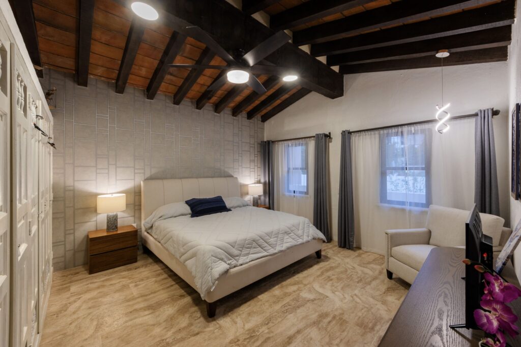 Bedroom with a tufted beige bed, two wooden nightstands, and a ceiling fan under dark wooden beams; light gray wall panels and windows with sheer gray curtains on the right.