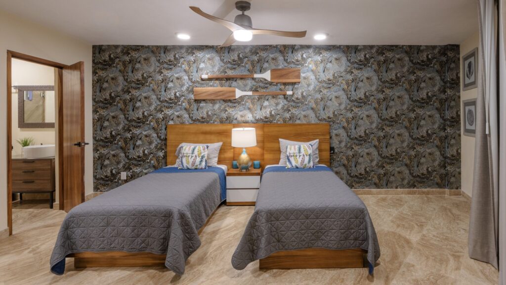 Twin beds with gray quilts and a shared wooden headboard in a room featuring a textured dark wall with wall-mounted oars as decor.