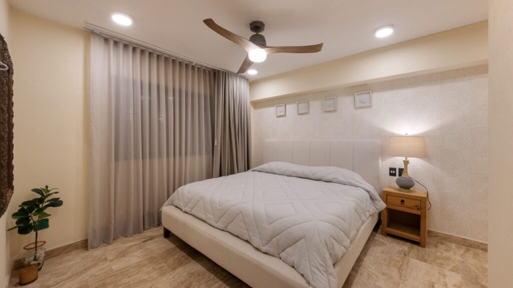 King bed with white quilt and upholstered headboard, wooden nightstand and lamp in a softly lit bedroom.