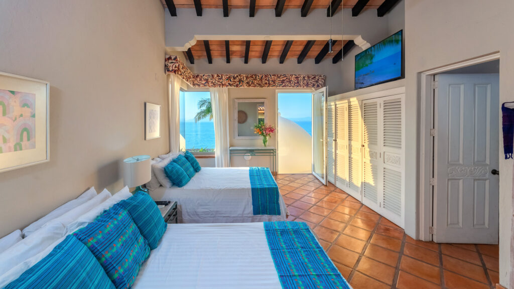Hotel room with white beds and blue bed runners, open balcony doors reveal blue ocean and palm tree outside, tropical decor.