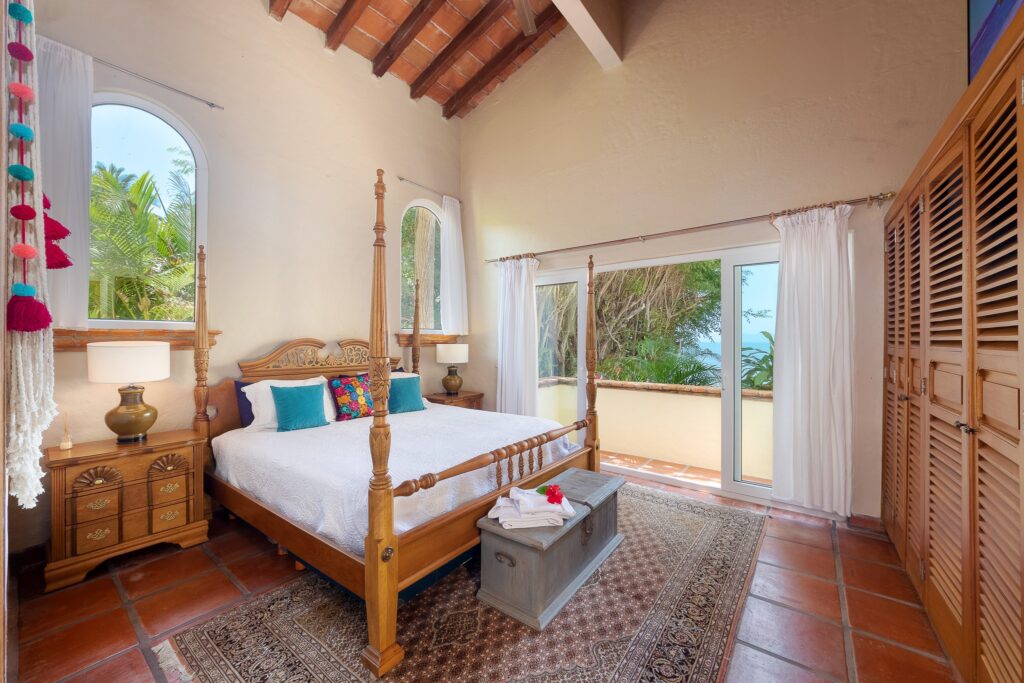 A sunlit tropical bedroom with a carved wooden four-poster bed, white bedding, colorful pillows, and two nightstands with lamps.