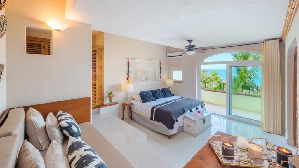 Bright coastal bedroom with a bed facing a balcony door overlooking palm trees and the sea