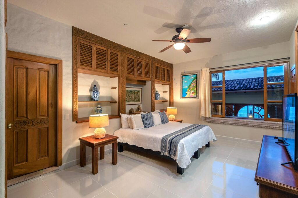 Bedroom with carved wooden shelves and cabinets above the bed, simple white bedding, and blue accents by two bedside lamps.