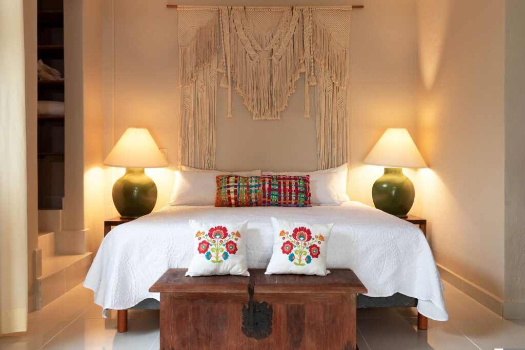 Bed with white quilt, colorful woven pillows, and a macramé wall hanging above; warm lamp light on both sides.