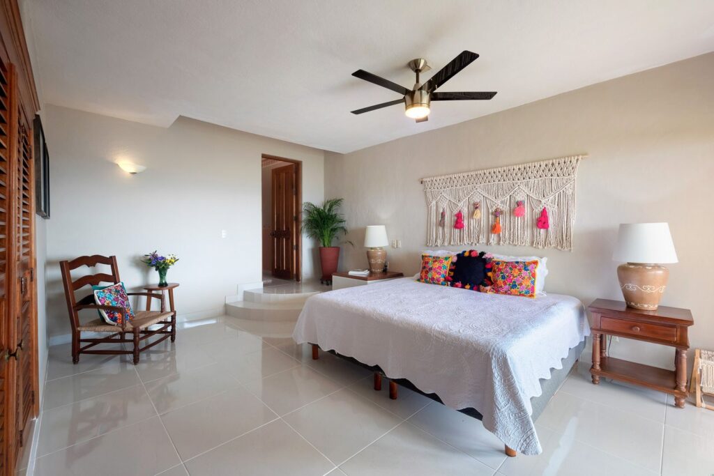 Queen bed with white quilt and colorful pillows, macrame wall hanging, two nightstands, and a ceiling fan in a bright bedroom.