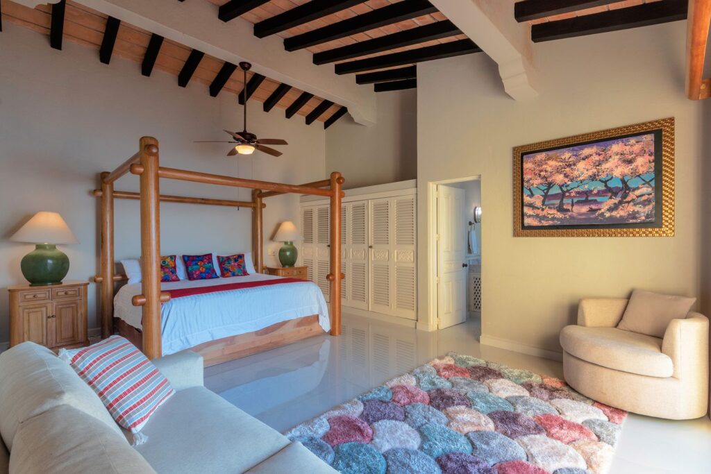 Large wooden four‑poster bed with blue bedding and colorful pillows, flanked by matching green lamps in a bright, airy bedroom.