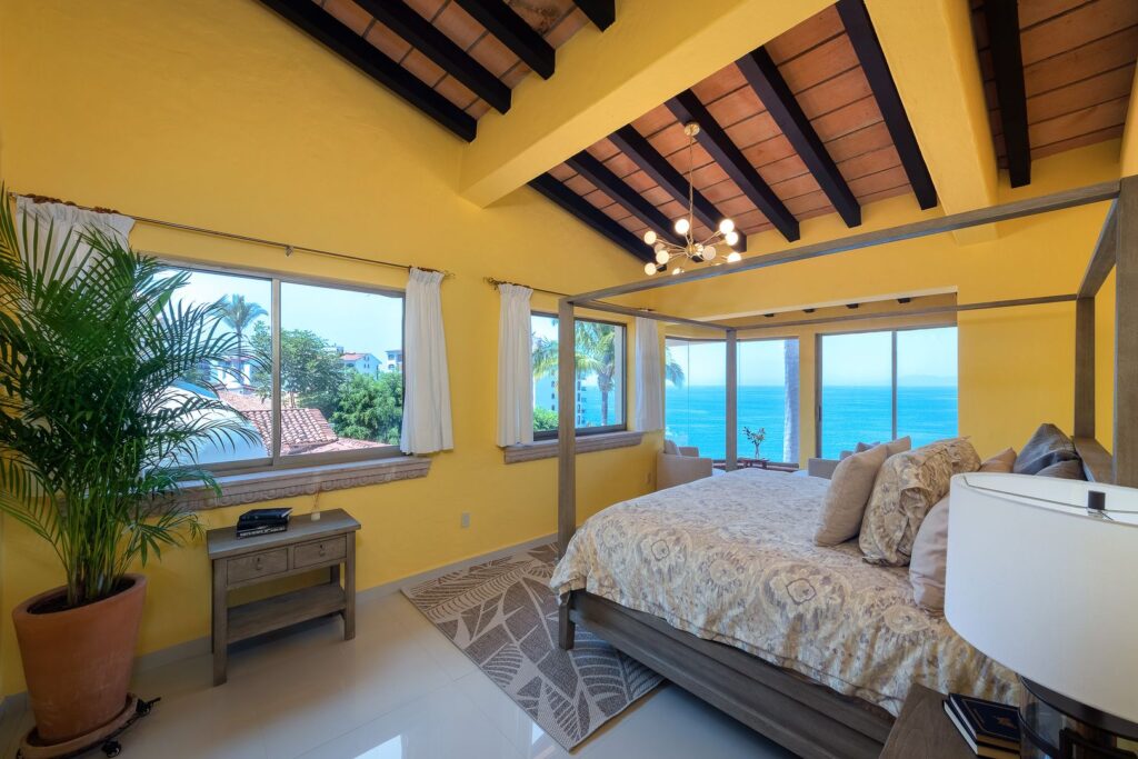 Bright yellow bedroom with a four-poster bed, white bedding, and a view of the ocean through large windows; a potted palm sits by the wall.