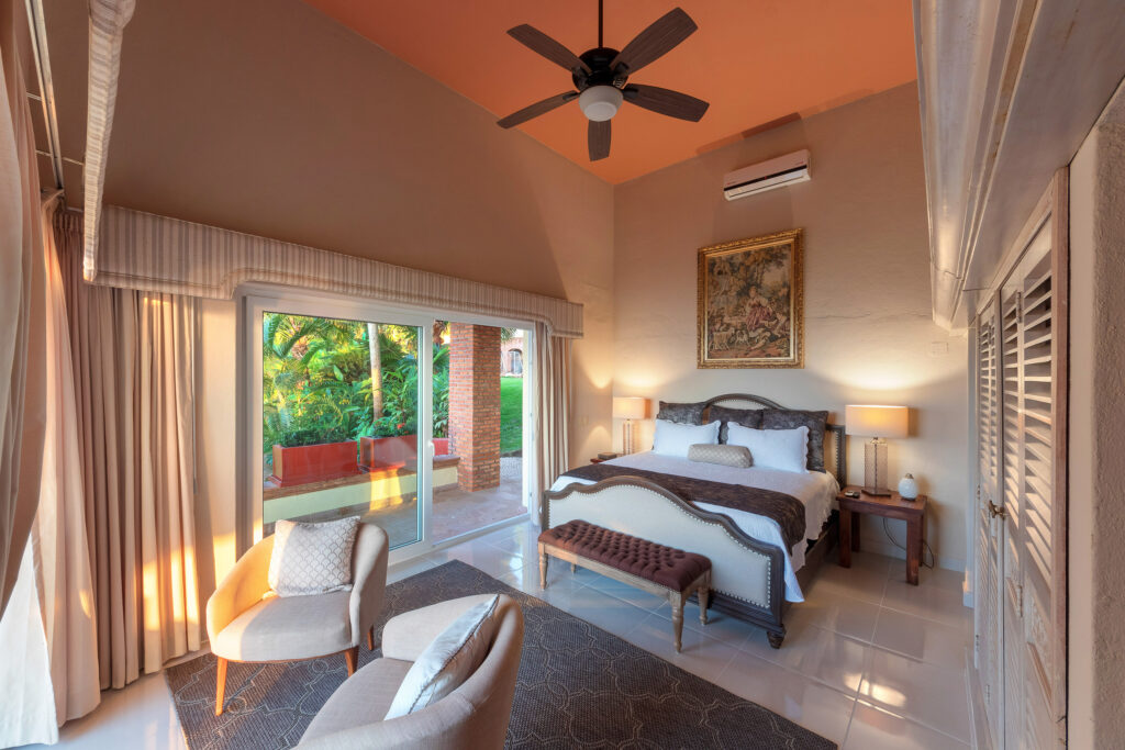Hotel bedroom with two matching beds, a bench at the foot, and a seating area near a sliding glass door to a patio garden outside.