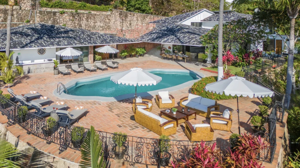 Poolside resort scene with a curved turquoise pool, white umbrellas, and sun loungers on terracotta tiles.