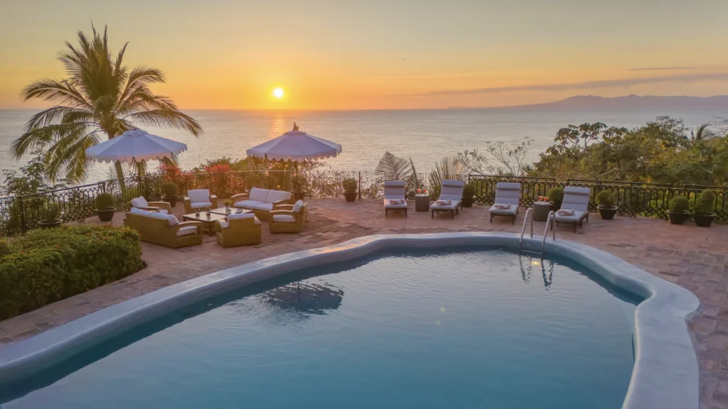 Poolside terrace at sunset overlooking the ocean with white umbrellas and cushioned seating.