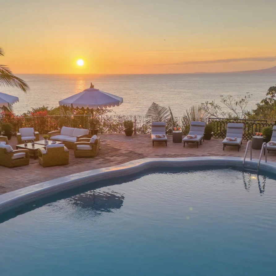 Poolside terrace at sunset overlooking the ocean with white umbrellas and cushioned seating.