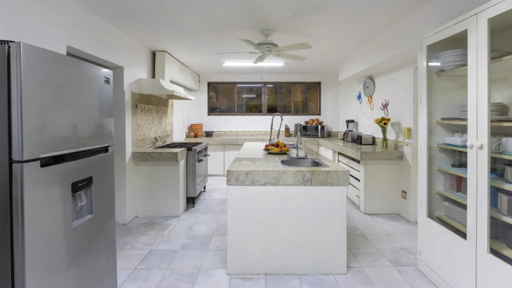 Bright U-shaped kitchen with a center island, stainless fridge, and granite countertops. Windowed wall at back, ceiling fan overhead.