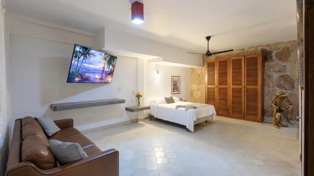 Hotel room featuring a queen bed, brown leather sofa, wall-mounted TV, and a stone wall with wooden closet doors.