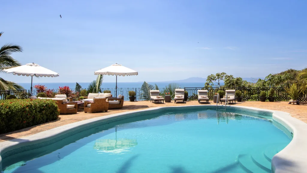 Poolside terrace with white umbrellas, wicker seating, and an ocean view beyond a decorative railing on a sunny day.