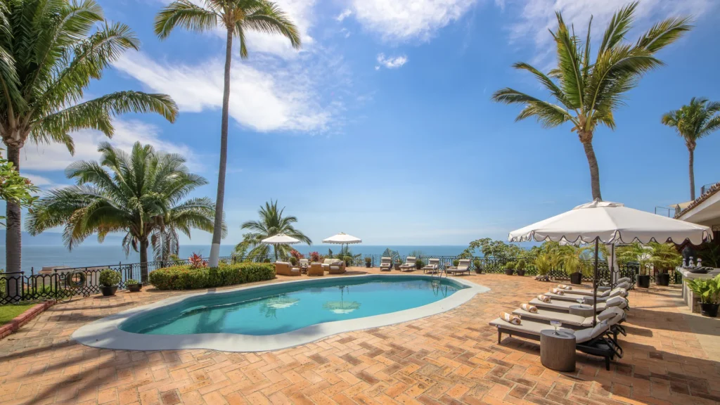 Poolside at a tropical resort: palm trees, white umbrellas, sun loungers, and a sea view.