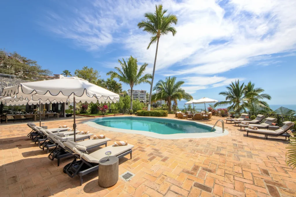Resort pool area with sun loungers under umbrellas, palm trees, and ocean view in the distance in bright sunlight.