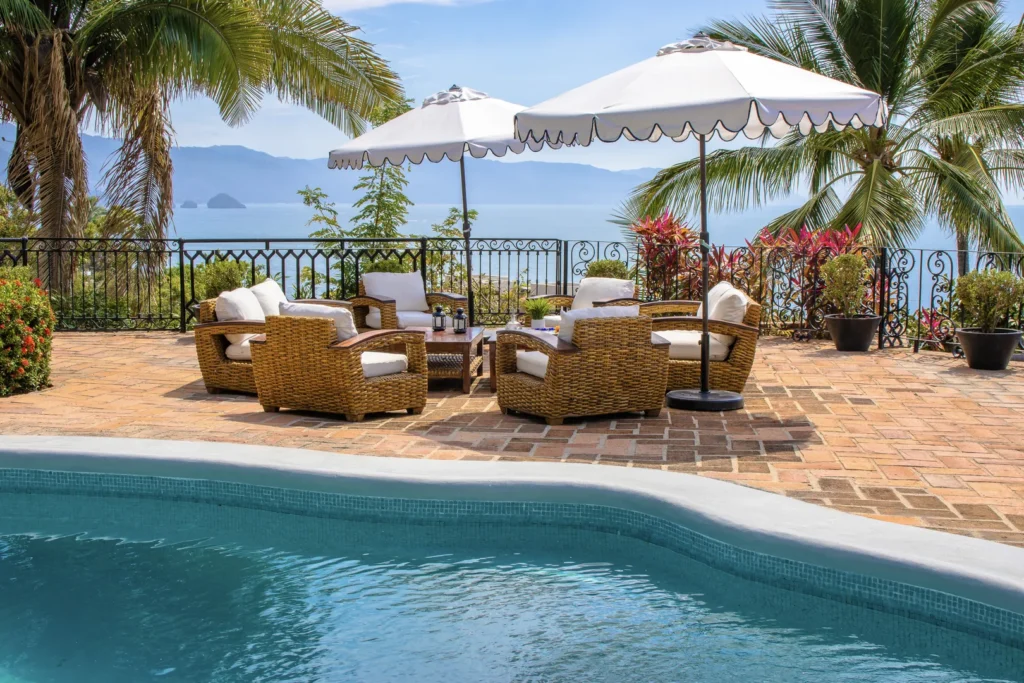 Wicker outdoor seating area by a swimming pool with white cushions and two white umbrellas, overlooking the sea and palm trees.
