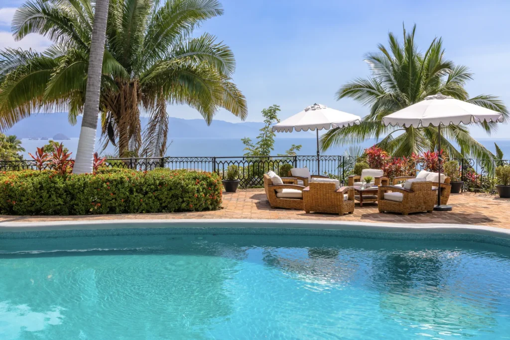 Poolside patio with wicker lounge chairs under white umbrellas, tropical plants, and a sea view