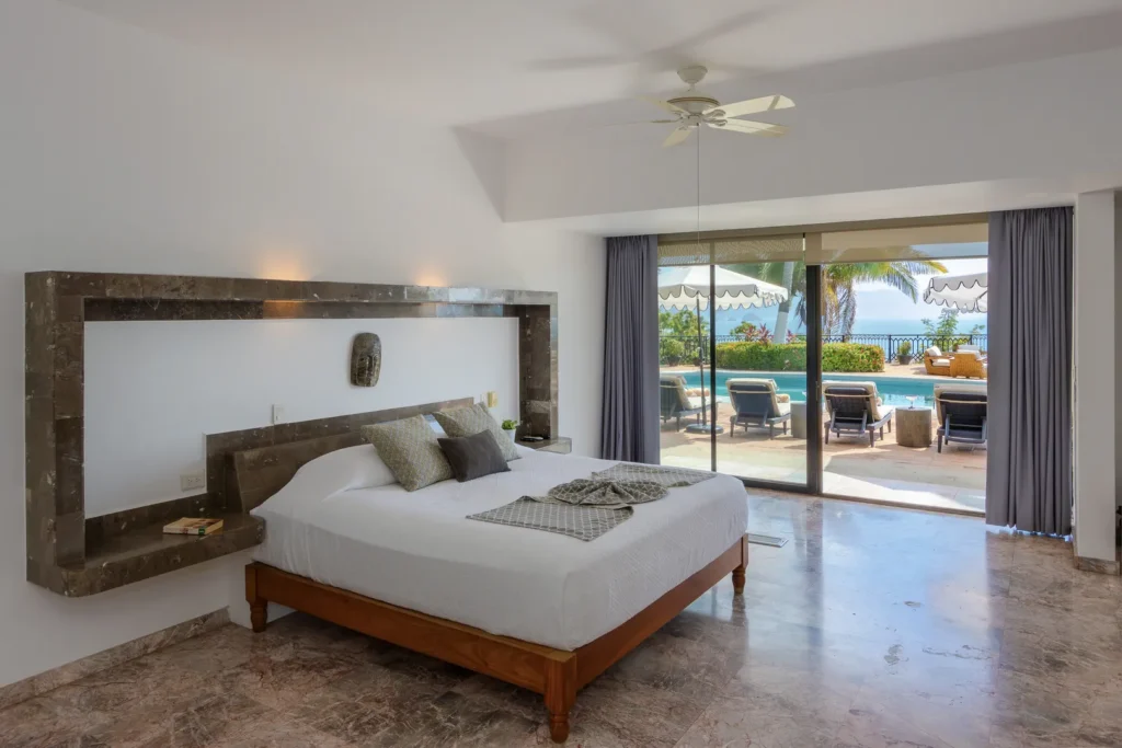 Bedroom with a king bed, marble-framed headboard, and sliding glass doors opening to a pool and ocean view beyond.