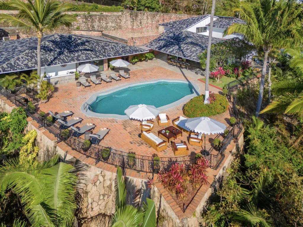 Aerial view of a resort pool area with curved turquoise pool, white umbrellas, and loungers on a terracotta patio amid tropical plants and stone walls,