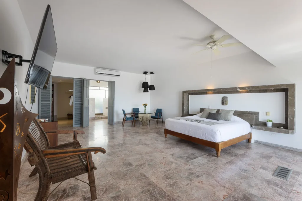 Spacious hotel suite with a king bed, marble floor, and a seating area of blue chairs around a glass table near the doorway.