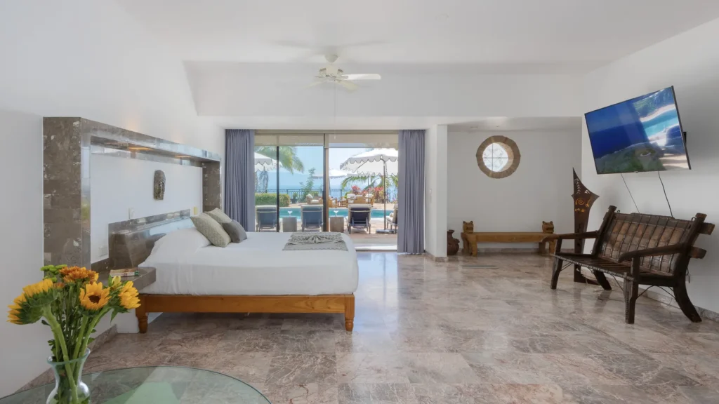 Bright hotel suite with a large bed, marble floor, and a sliding glass door opening to a pool area and ocean view beyond.