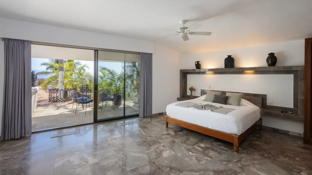 Hotel bedroom with a king bed, white bedding, and marble floors; sliding doors open to a patio with chairs and palm trees outside.