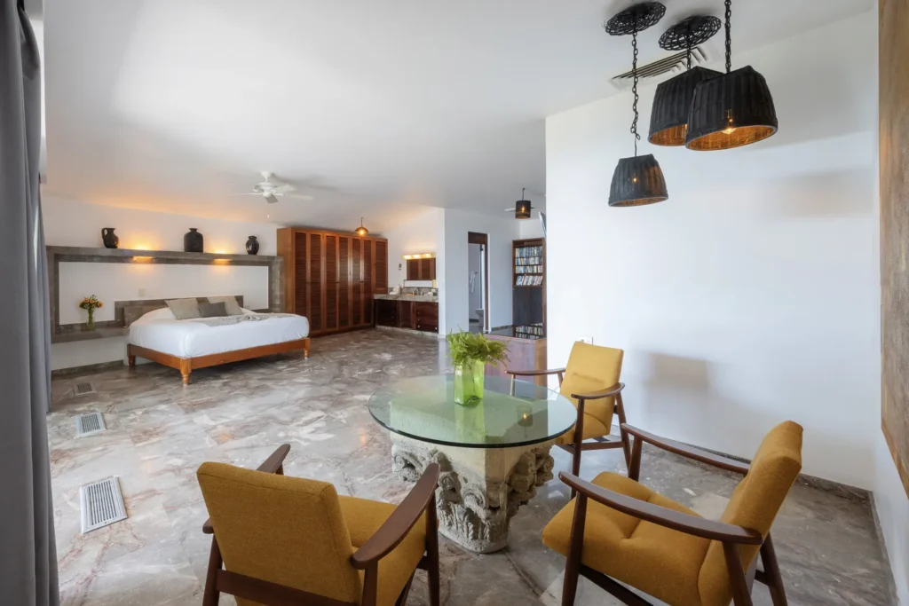 Hotel suite living area with a bed, marble floor, and a glass round table surrounded by mustard chairs.