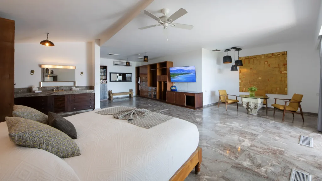 Spacious hotel suite with a large bed in the foreground, marble floors, and a TV lounge area.