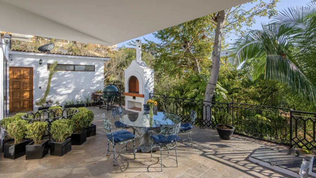 Sunny outdoor patio with a glass round table, blue-cushioned metal chairs, and a white outdoor oven, surrounded by potted plants and tropical trees.