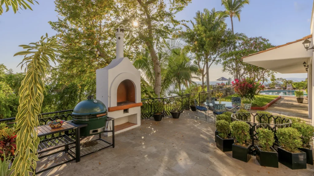 Sunlit outdoor patio with a white brick oven, a green ceramic grill, a glass table, and potted plants by a pool. In the distance, tropical trees frame the scene.
