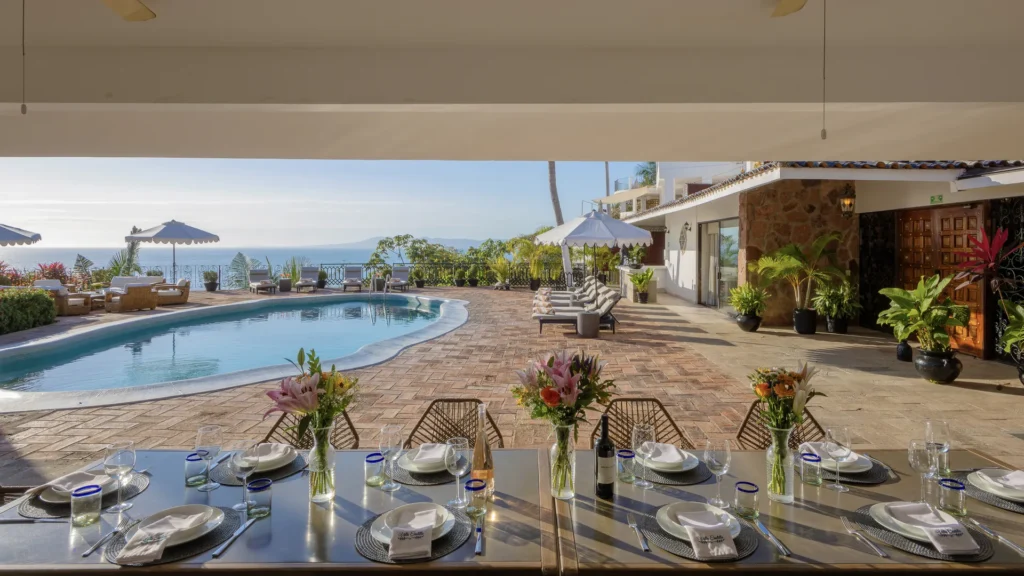 Poolside dining table set for a meal on a sunny terrace overlooking the ocean, with lounge chairs and umbrellas in the background.