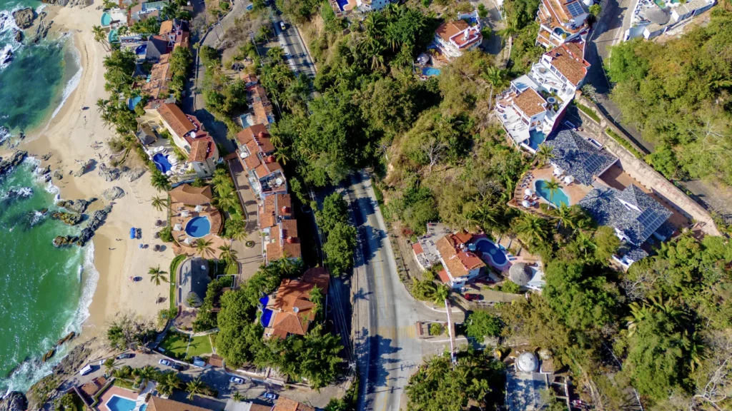 Aerial view of a beachfront residential area with red-tiled roofs, palm trees, and a central tree-lined road running through the neighborhood.