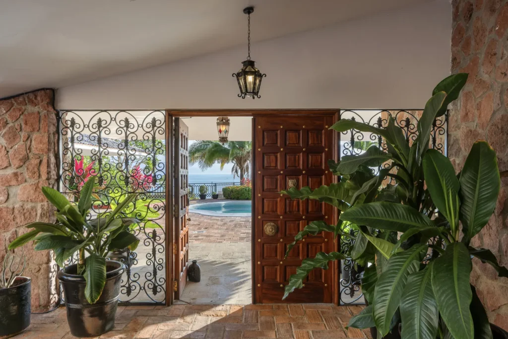Open wooden door with brass studs opens to a sunlit courtyard, pool and ocean beyond, framed by wrought-iron gates and potted plants.