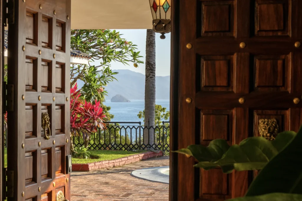 Open wooden doors frame a tropical garden and ocean view with a palm tree and distant islands beyond.