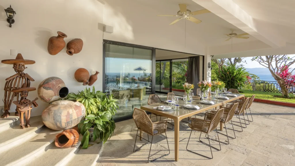 Outdoor dining area with a long table set for a meal, wicker chairs, and a sea view beyond glass doors.