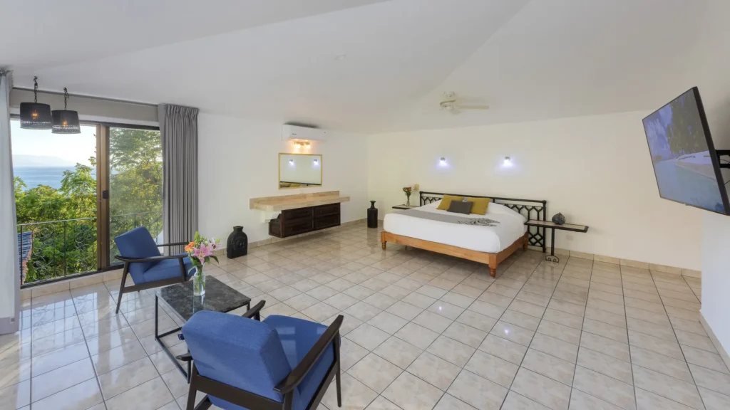 Spacious hotel room with a large bed, blue lounge chairs, and a balcony with an ocean view beyond the railing.