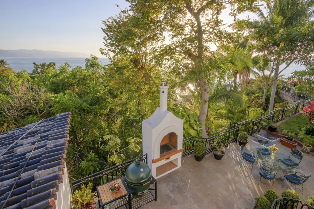Terrace with a white outdoor oven, glass table and blue chairs, overlooking lush greenery and the ocean beyond as the sun shines through trees.
