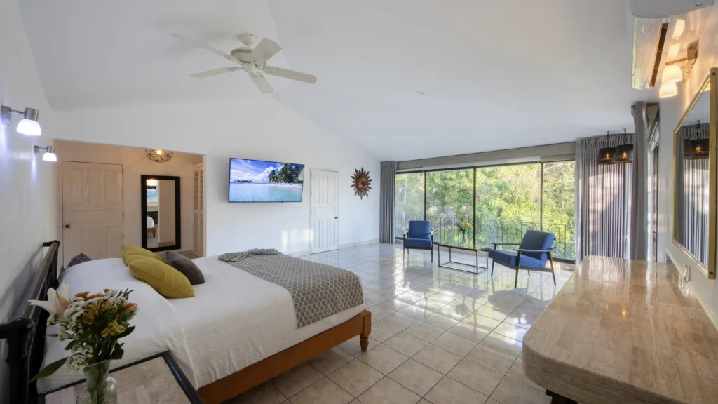 Spacious hotel suite with a large bed, wall-mounted TV, and a seating area by glass doors opening to a balcony.